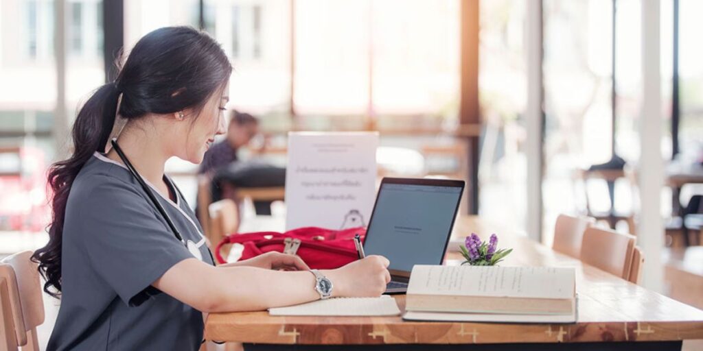  A young nurse in professional scrubs with a stethoscope around her neck is sitting at a wooden table in a bright, modern workspace. She is writing notes on paper while working on a laptop, preparing her resume for job applications.